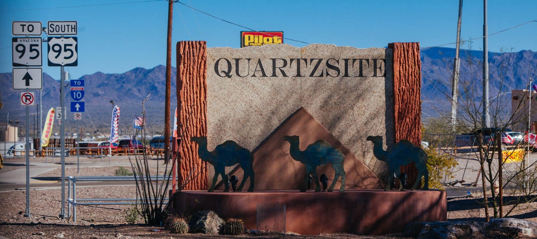 A stone entrance sign for Quartzsite, Arizona, adorned with metal camel silhouettes, surrounded by desert scenery and road signs
