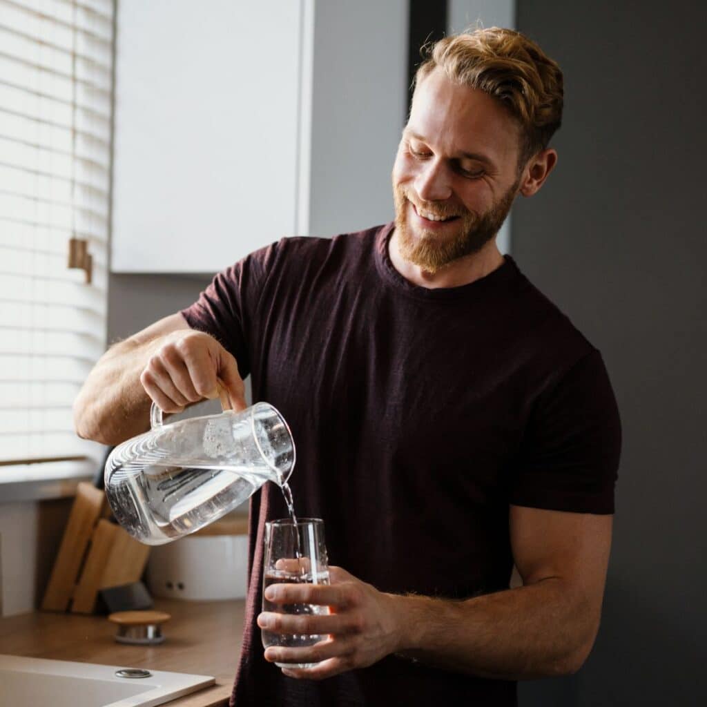A smiling man pours water from a glass pitcher into a drinking glass at the kitchen sink