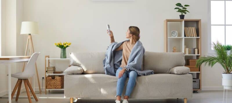 Woman sitting on a couch with a blanket, pointing a remote at the air conditioner in a bright living room.